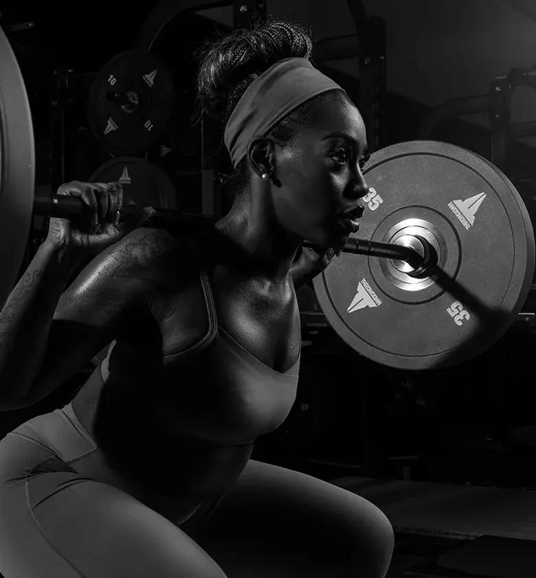 Black-and-white photo of a woman in a headband and sports bra squatting with a barbell across her shoulders, loaded with Throwdown 35 lb bumper plates, in a dimly lit gym.